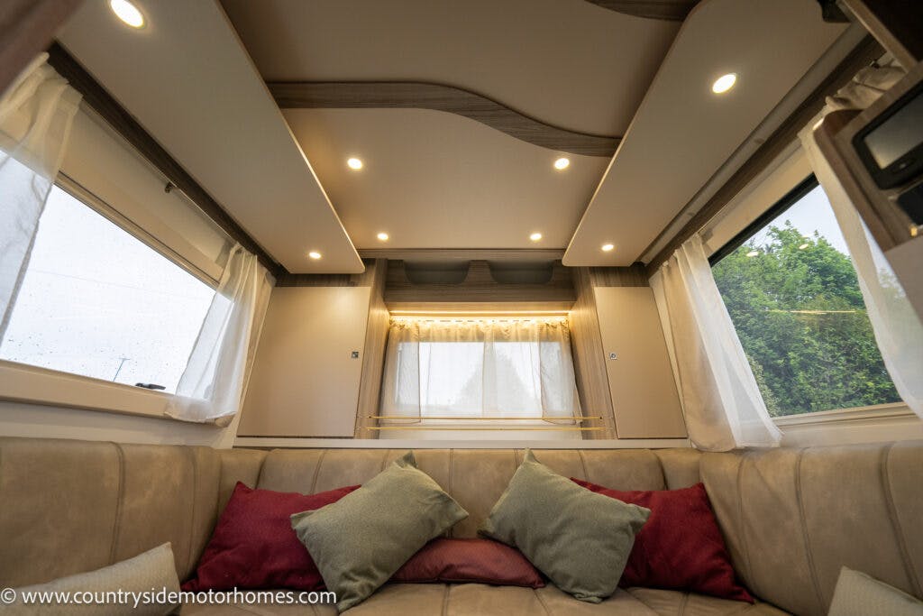 The interior of the 2022 Benimar Mileo 282 motorhome features beige and maroon cushions on a U-shaped seating area. Windows with white curtains are on three sides, and recessed lighting is installed in the ceiling. Countrysidemotorhomes.com is visible in the bottom left corner.