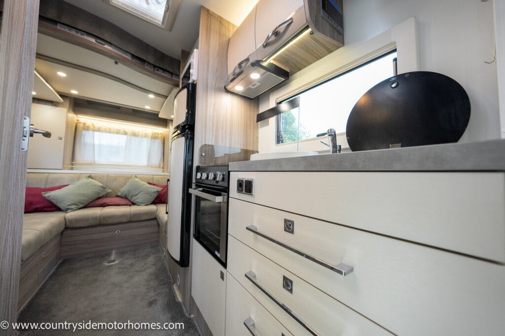 Interior of the 2022 Benimar Mileo 282 motorhome featuring a kitchen area with white cabinets, an oven, and a stove. The left side has a seating area with a beige sofa and colorful cushions. A window above the sink allows natural light in. The URL www.countrysidemotorhomes.com is visible.