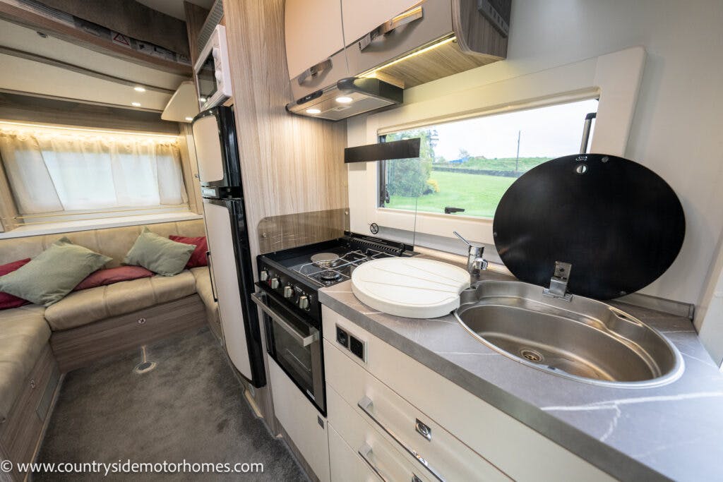 The image shows the interior of a 2022 Benimar Mileo 282 motorhome kitchen area with a sink, a stove, an oven, and overhead cabinets. There is a window above the countertop. In the background, there's a living area with a beige sofa and throw pillows.