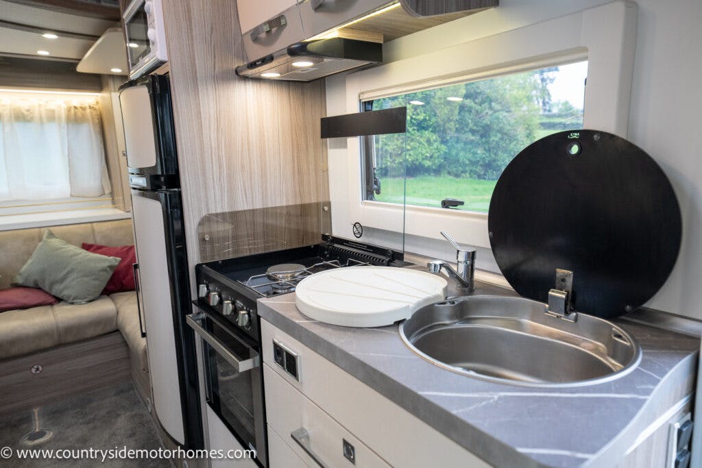 The interior of the 2022 Benimar Mileo 282 motorhome kitchen features a compact sink, a stove with an oven, and overhead storage cabinets. Adjacent to the kitchen area is a small seating space with a sofa and pillows. A window is situated above the kitchen counter.