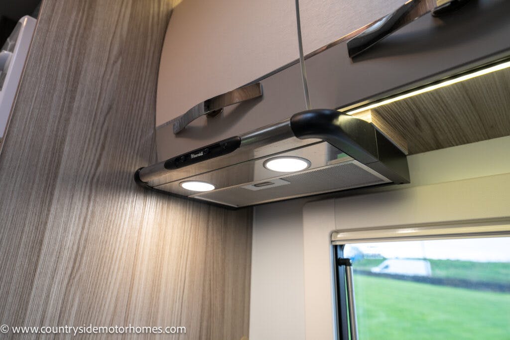 A close-up view of a modern kitchen range hood with two lit LED lights beneath a cabinet in the 2022 Benimar Mileo 282. The range hood has metallic accents and is installed above a cooktop. A window is partially visible in the background, letting in some natural light.