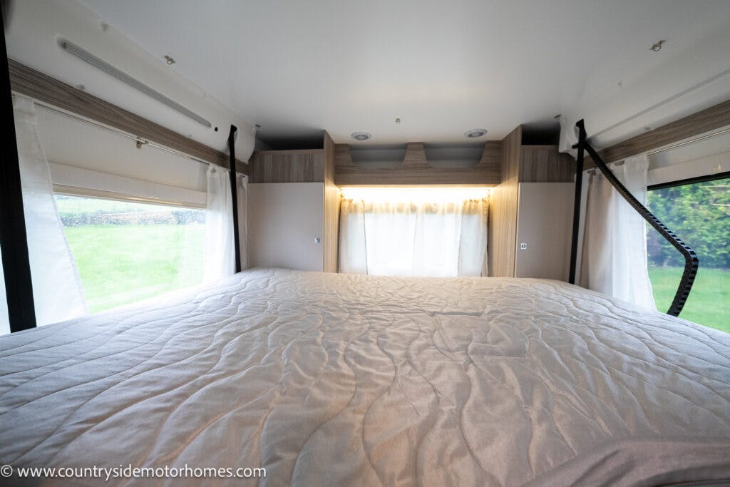 This image shows the interior of a 2022 Benimar Mileo 282 motorhome bedroom with a large, neatly made bed covered in a light-colored quilt. The room features wooden accents and large windows with curtains that allow plenty of natural light to enter. There is greenery visible outside.