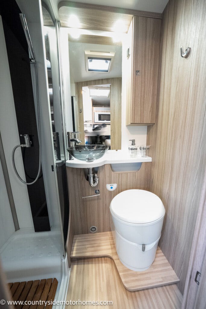 The image shows the interior of a 2022 Benimar Mileo 282's bathroom. It features a white toilet, a vessel sink with chrome faucet, narrow countertop, and mirrored cabinets. There is a shower area to the left with a frosted glass door. The walls are paneled with wood-like material.