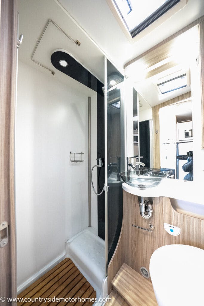 This image shows the interior of a 2022 Benimar Mileo 282 motorhome bathroom. The space includes a shower area with a glass door, a sink with a faucet, a mirror, a wooden floor mat, and various storage compartments. The overall design is modern with light wood and white finishes.