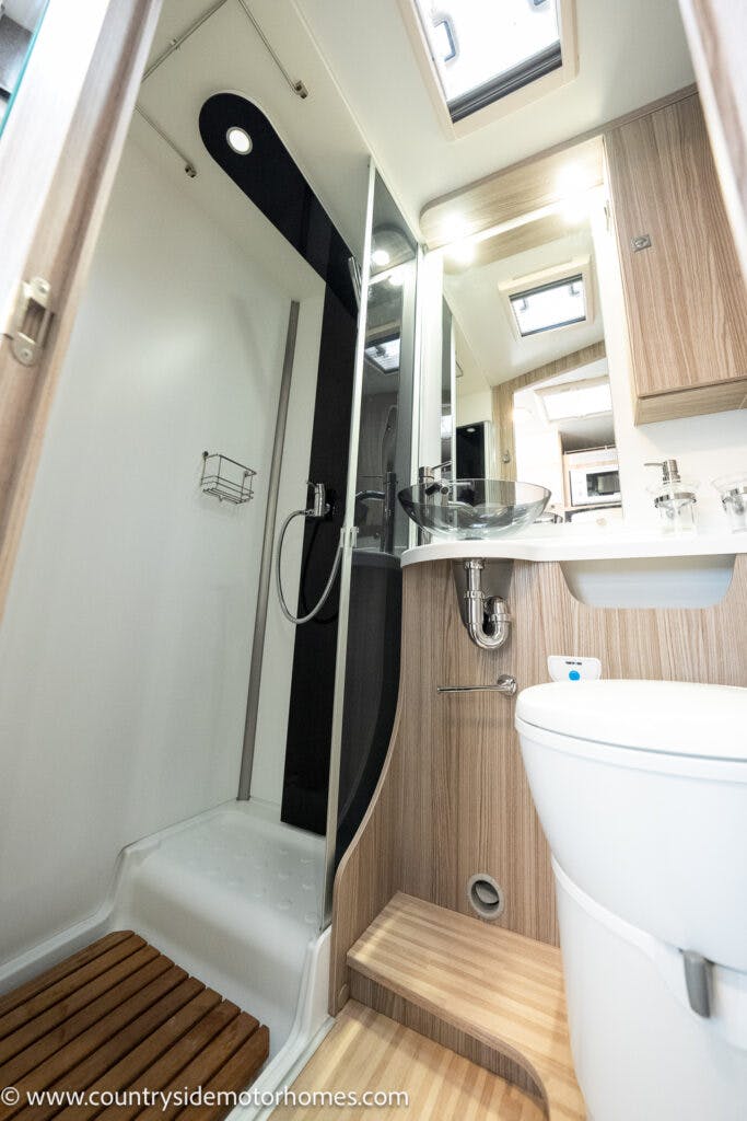 This image shows the interior of a 2022 Benimar Mileo 282 motorhome bathroom. It features a shower area with a glass door, a sink with a mirror above it, and a toilet. Wooden accents and built-in storage space are visible. An overhead skylight provides natural light.