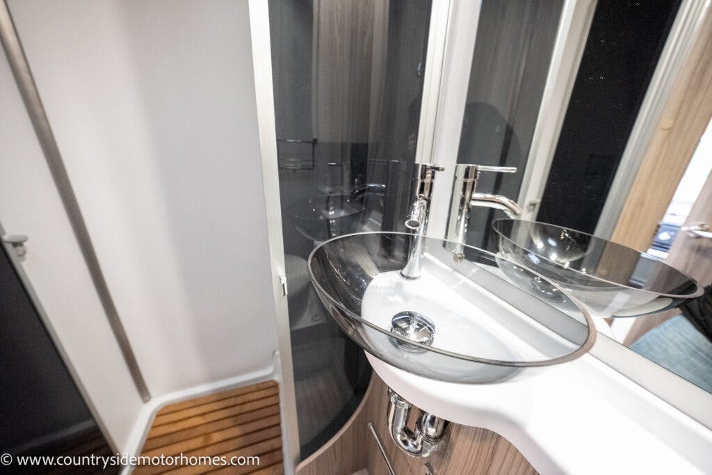 A compact bathroom in the 2022 Benimar Mileo 282 RV features a modern glass vessel sink with a chrome faucet set on a white counter. A glass-enclosed shower area is visible in the background. The floor has a wooden slat design. The image is sourced from countrysidemotorhomes.com.