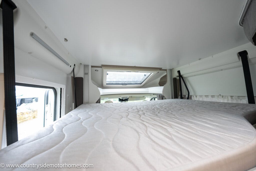 The interior of the 2022 Benimar Mileo 282 motorhome is shown, featuring a bed area with a large mattress and a skylight overhead. Straps and fixtures are visible on the walls, indicating storage or securing mechanisms, and a website URL is visible at the bottom left corner.