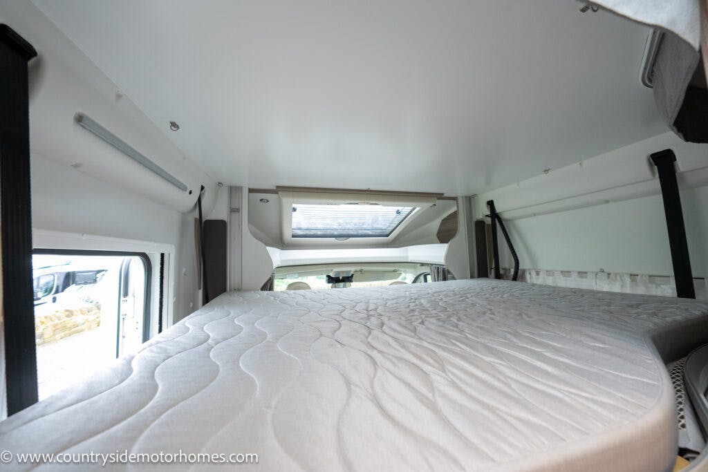The image shows the interior of a 2022 Benimar Mileo 282 motorhome viewed from the rear. The main focus is on a raised bed area with a mattress. There is a skylight window above, and the surrounding walls are white. A URL, countryside motorhomes dot com, is visible in the lower left corner.