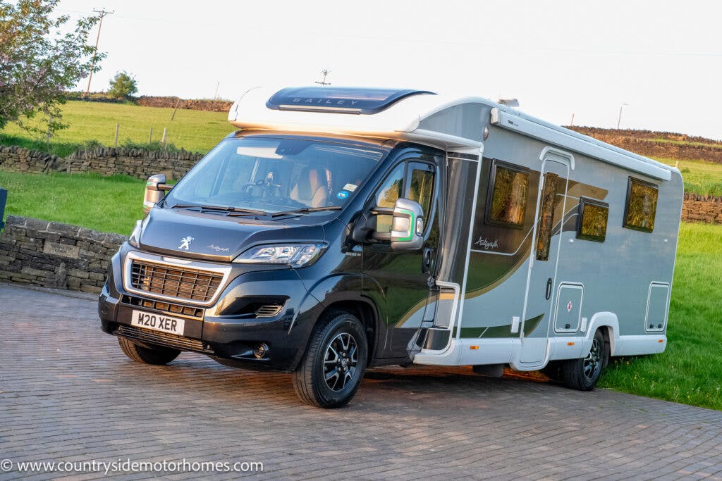 A modern 2021 Bailey Autograph 79-4i motorhome with the license plate "M20 XER" is parked on a paved area. The vehicle has a black and silver color scheme and appears to be well-maintained. There is a grassy field and stone wall in the background. The website countryside motorhomes is visible.