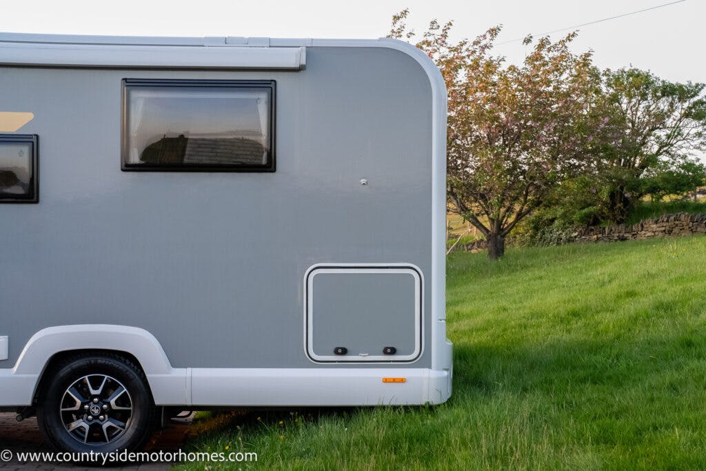 Side view of a grey 2021 Bailey Autograph 79-4i motorhome parked on a grassy area with a tree and stone wall in the background. The motorhome has a window, a hatch, and a tire visible. The image includes a visible website URL in the lower left corner.