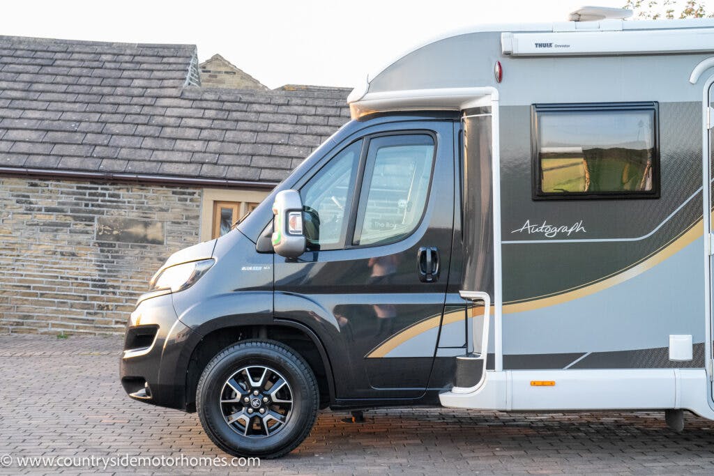 A gray motorhome, the 2021 Bailey Autograph 79-4i, is parked on a brick driveway. The motorhome features black wheels and a small window. In the background, you can see a brick building with a tiled roof.