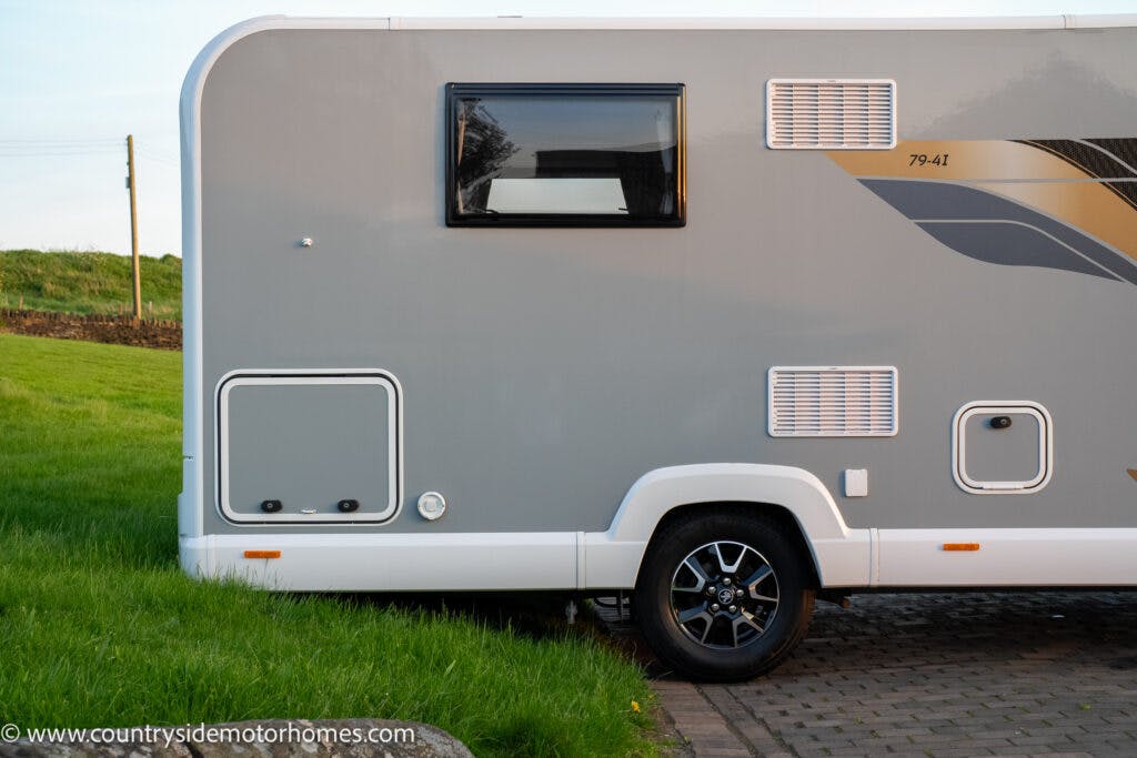 Side view of the 2021 Bailey Autograph 79-4i motorhome parked on a paved area next to a grassy field. The vehicle features a black-tinted window, multiple vents, and a single wheel with a stylish alloy rim. The countryside and utility poles are visible in the background.