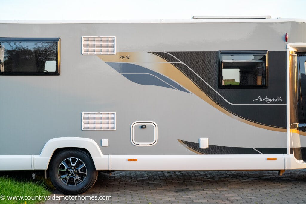 Side view of a grey 2021 Bailey Autograph 79-4i parked on a paved surface. The motorhome features blue and yellow wave patterns with the label "79-4T." Visible are windows and ventilation grilles. The website "countrysidemotorhomes.com" is written at the bottom.