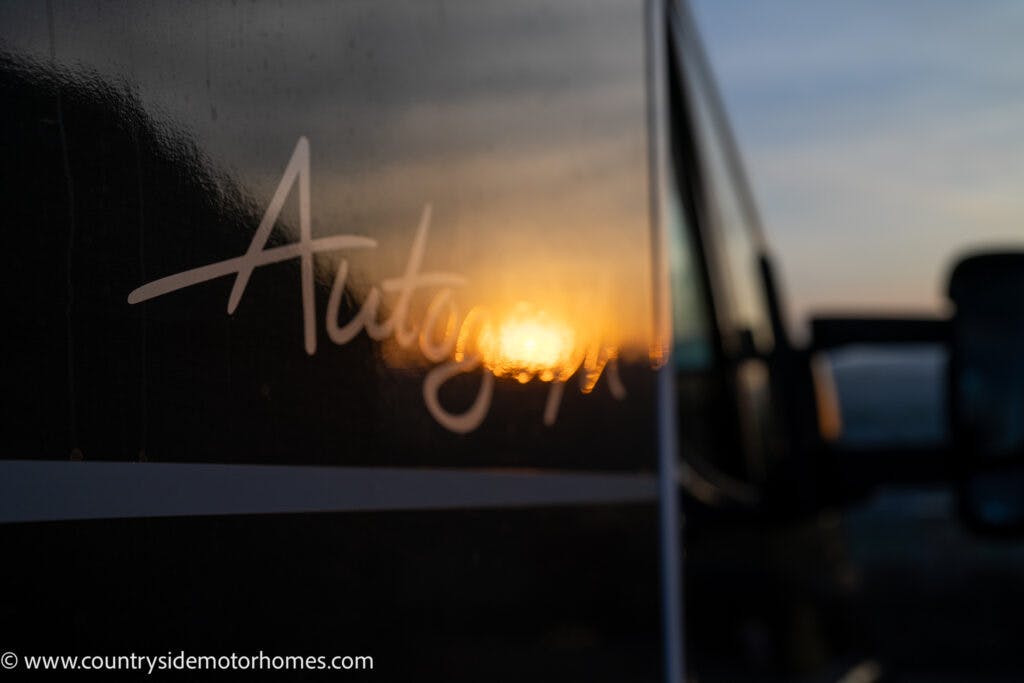 Close-up of a 2021 Bailey Autograph 79-4i motorhome with the word "Autograph" written on its side. The sunset is reflected on its glossy surface. The background is blurred, drawing focus to the reflection and the text. The website www.countrysidemotorhomes.com is visible.