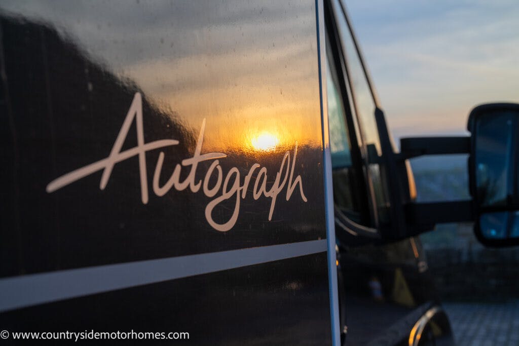 Close-up of a 2021 Bailey Autograph 79-4i motorhome with the name "Autograph" elegantly inscribed on its side, reflecting the golden hues of sunset. The dark body contrasts beautifully with the surrounding blurred outdoor setting, where a paved surface meets the distant horizon. A side mirror is also visible.