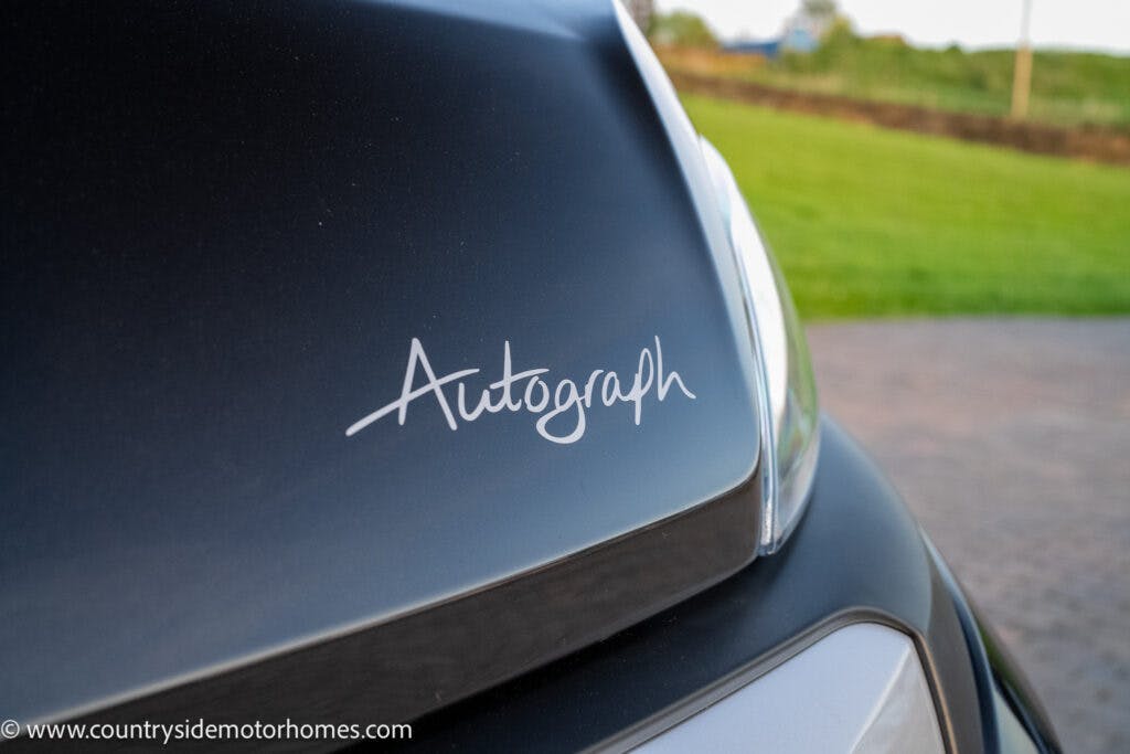 Close-up view of a 2021 Bailey Autograph 79-4i displaying the word "Autograph" in scripted font on its front. The foreground features part of the car's headlight and hood, with a blurred grassy field in the background. Website text "www.countrysidemotorhomes.com" is visible at the bottom left.