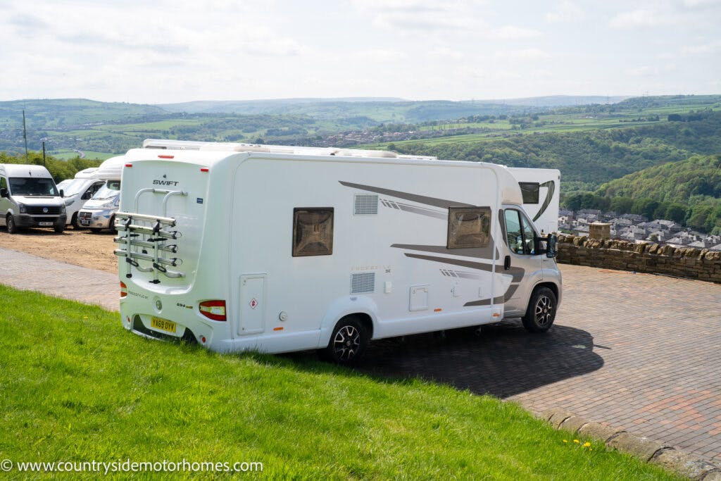 A 2019 Swift Escape 694 Freestyle motorhome is parked on a paved area overlooking a scenic, hilly landscape with green fields. There are other motorhomes parked in the background, and the sky is partly cloudy. A website URL, www.countrysidemotorhomes.com, is visible in the bottom left corner.