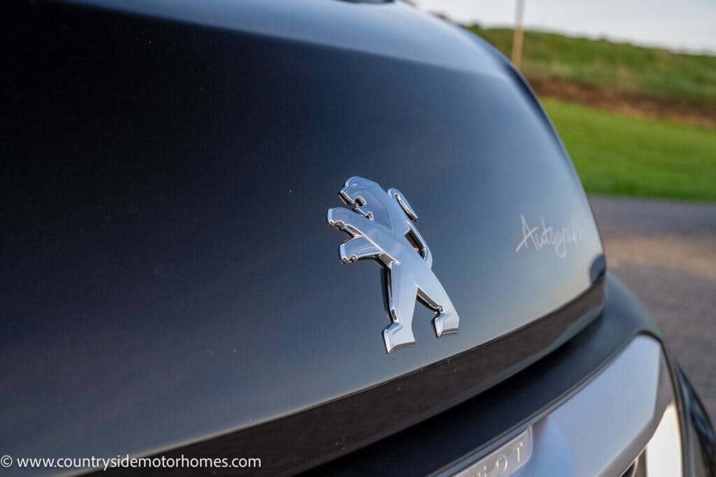 Close-up of a black vehicle's hood featuring a chrome Peugeot logo and the word "Autograph" on a glossy finish. The background shows a blurred landscape with green grass. The image, highlighting the 2021 Bailey Autograph 79-4i, includes a web address at the bottom left corner.