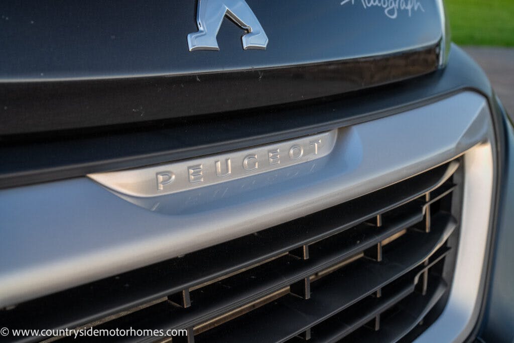 Close-up of the front grille of a vehicle featuring the Peugeot logo and brand name. The 2021 Bailey Autograph 79-4i emblem shines above the grille. The background includes some greenery, while the website URL is visible at the bottom left corner.