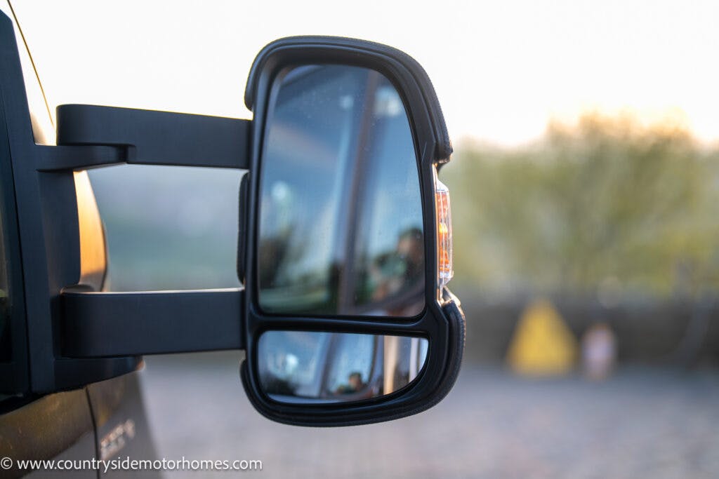 Close-up of a side mirror on a 2021 Bailey Autograph 79-4i motorhome, showing a sunlit outdoor scene reflected in the mirror. The background is blurred, including a triangular yellow sign and greenery. The motorhome's website URL is visible in the bottom left corner: www.countrysidemotorhomes.com.