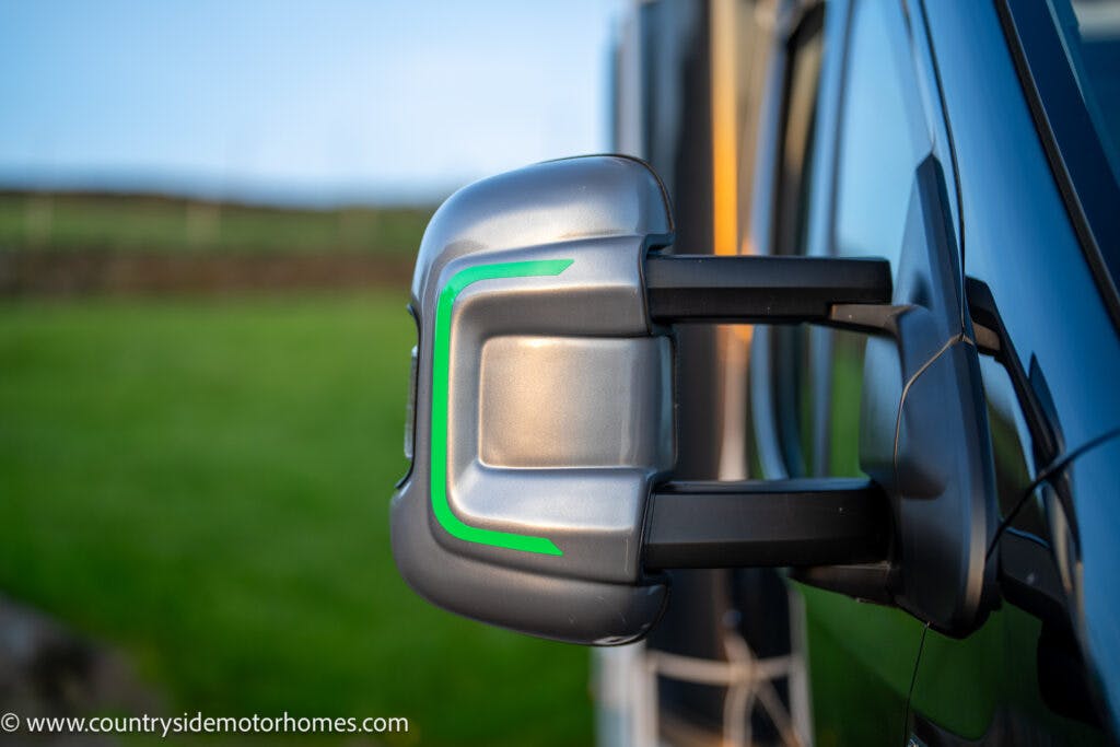 Close-up of a retracted 2021 Bailey Autograph 79-4i campervan mirror. The side mirror boasts a futuristic design with a silver casing and green stripe. In the background, a grassy field and blurred landscape are visible, along with a web address in the lower-left corner of the image.
