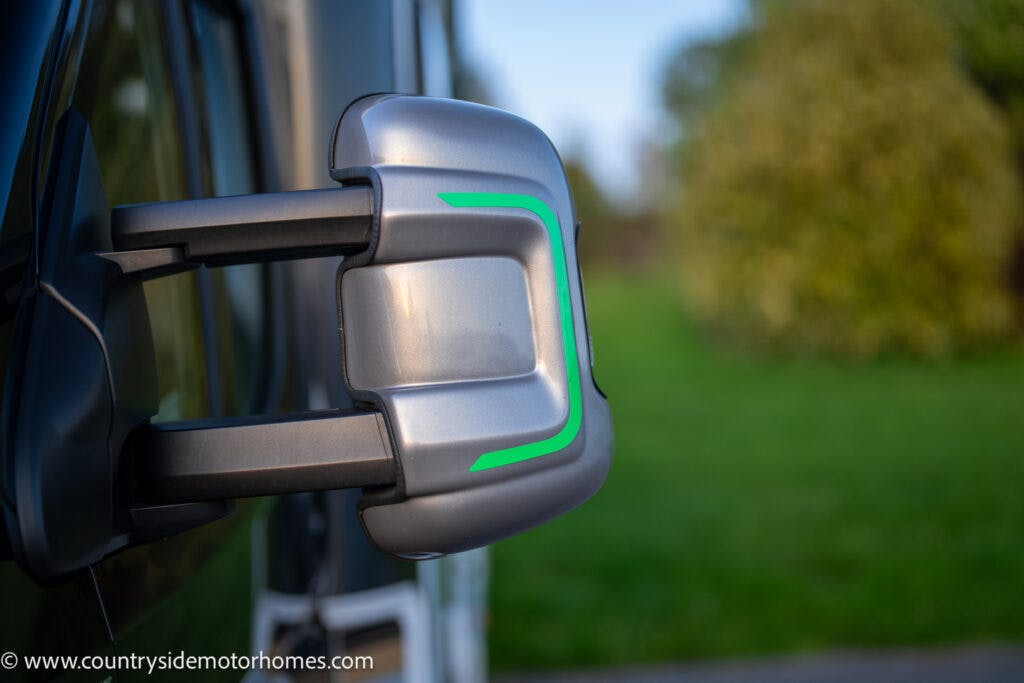 Close-up shot of a silver side mirror of the 2021 Bailey Autograph 79-4i motorhome with a green accent, facing a blurred green landscape. The website www.countrysidemotorhomes.com is visible in the bottom left corner.