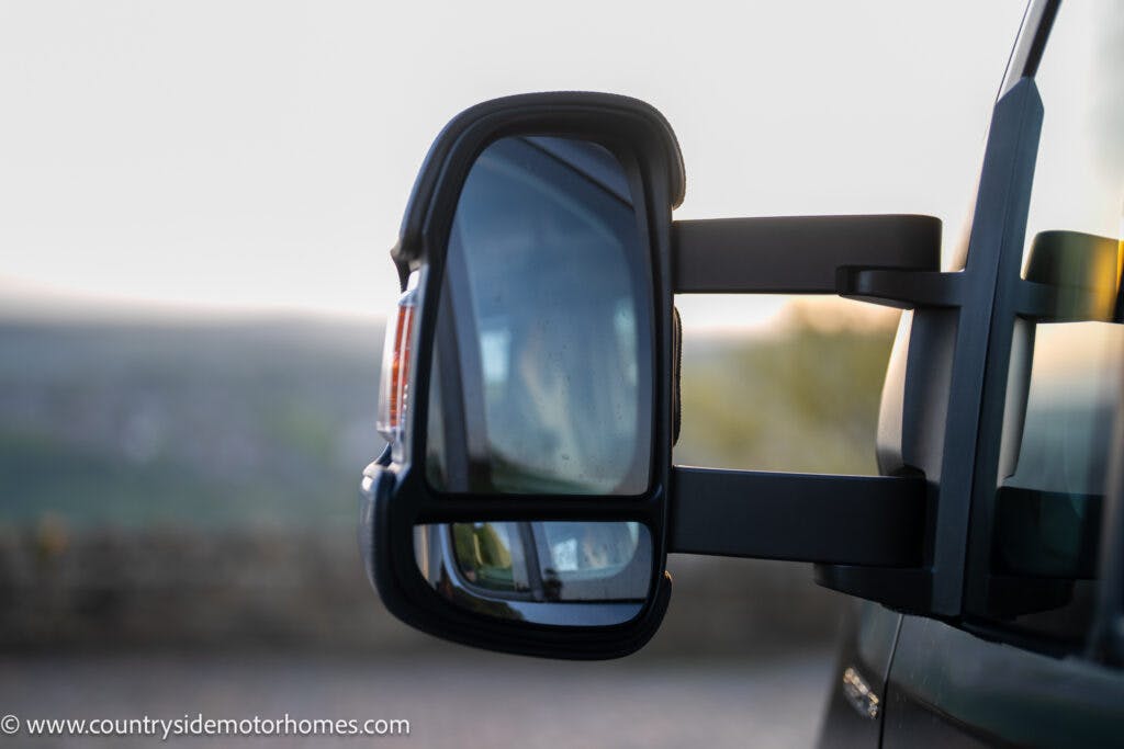 A close-up view of a side mirror on a 2021 Bailey Autograph 79-4i motorhome against a blurred background of countryside and sunset. The mirror reflects part of the motorhome and surroundings. The URL www.countrysidemotorhomes.com is visible in the bottom-left corner.