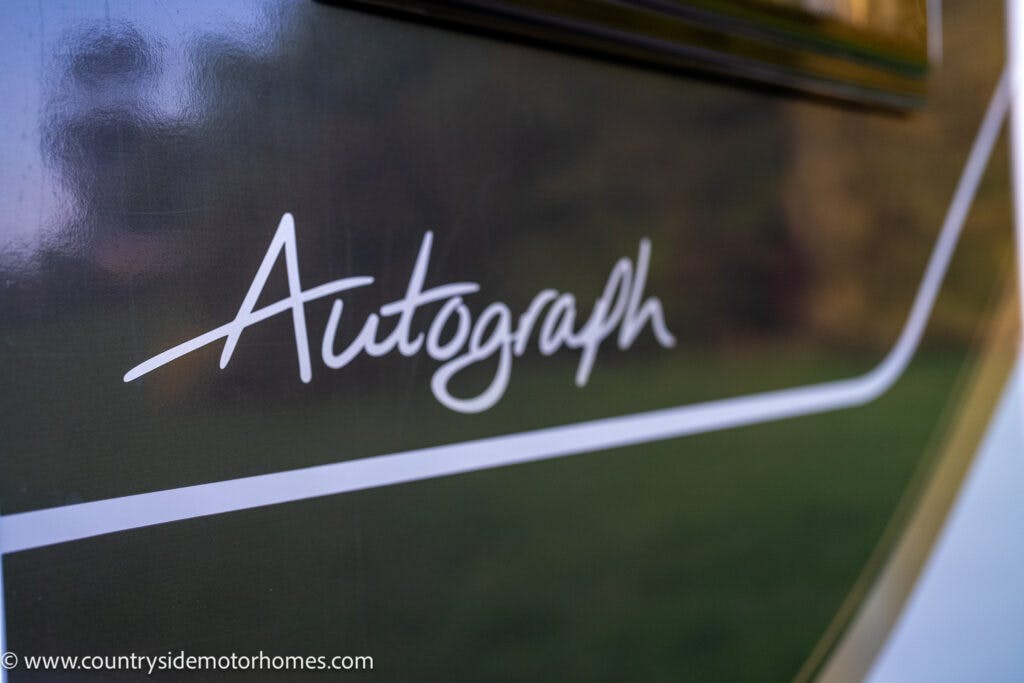Close-up of the word "Autograph" written in cursive on the side of a 2021 Bailey Autograph 79-4i with a dark background and a light diagonal stripe. The website address "www.countrysidemotorhomes.com" is visible in the lower left corner of the image.
