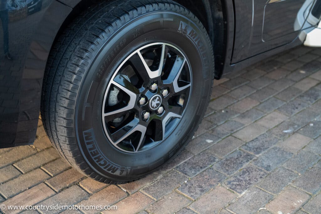 Close-up image of a 2021 Bailey Autograph 79-4i's front tire and wheel on a brick-paved surface. The Michelin tire is mounted on a six-spoke alloy wheel, with part of the car's body visible. A watermark in the corner reads www.countrysidemotorhomes.com.