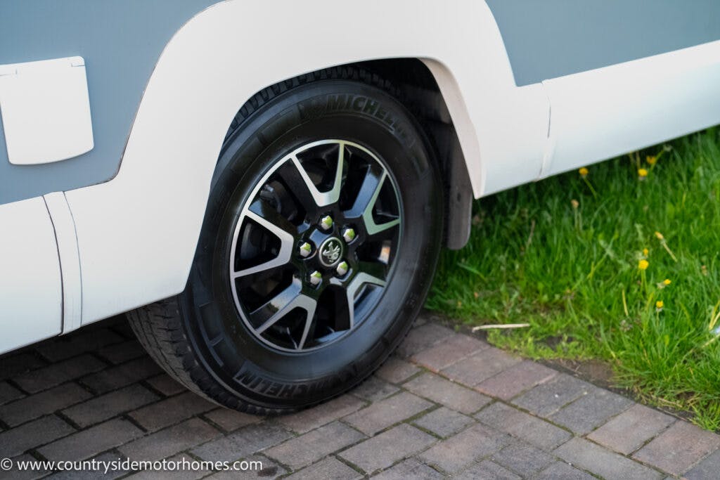 A close-up view of a 2021 Bailey Autograph 79-4i's rear tire and rim, showcasing the Michelin branding. The tire is mounted on a paved surface, with part of the grassy lawn visible adjacent to the pavement. The website "www.countrysidemotorhomes.com" is visible at the bottom left.