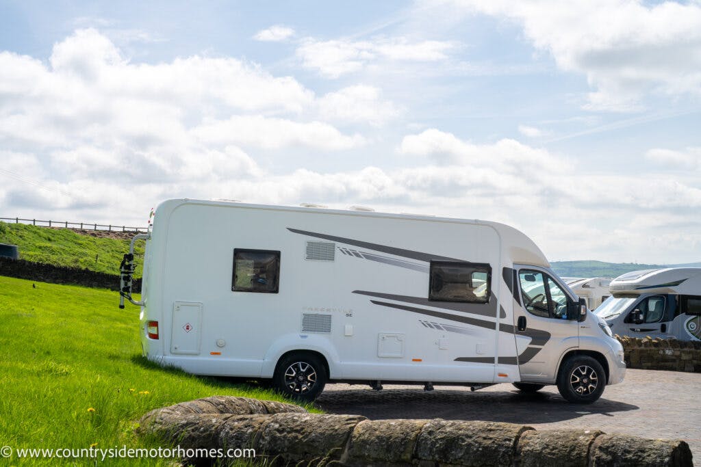 A white 2019 Swift Escape 694 Freestyle motorhome is parked on a paved area with a grassy field and stone wall nearby. The sky is partly cloudy, and another motorhome is visible in the background. A website URL for countrysidemotorhomes.com is displayed at the bottom left corner of the image.