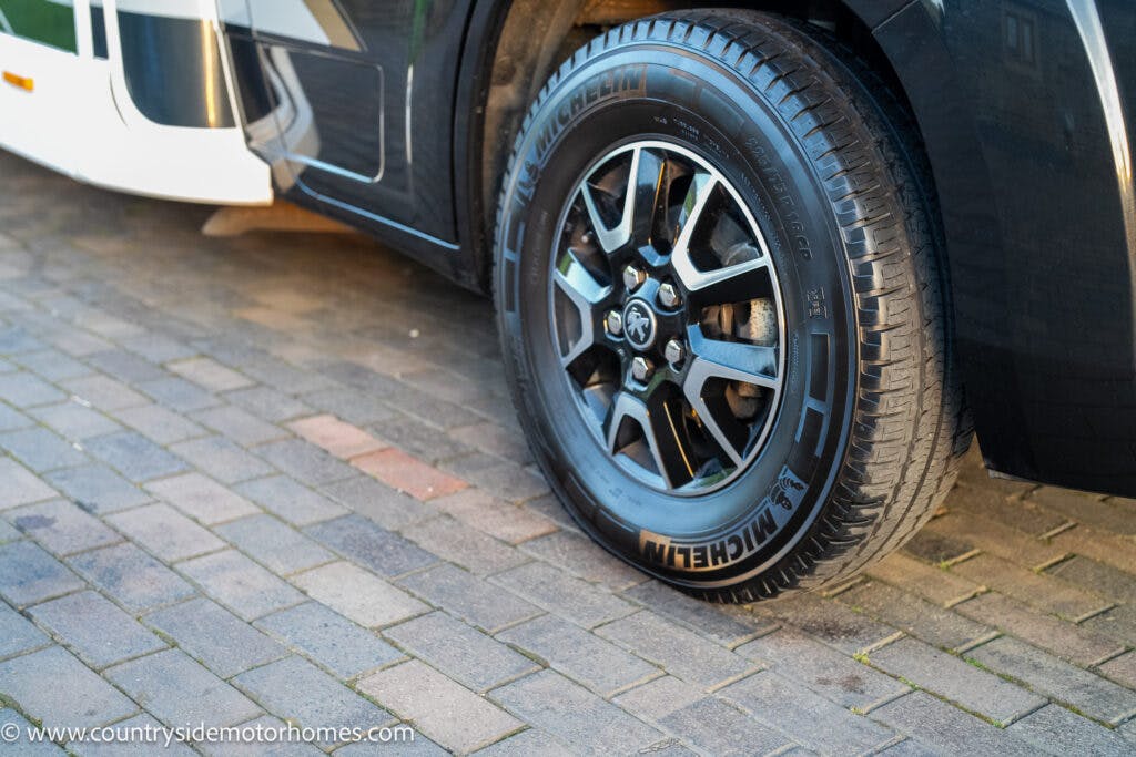 A close-up image captures the front tire of a black 2021 Bailey Autograph 79-4i motorhome parked on a brick driveway. The Michelin tire is mounted on a sleek black alloy rim, with part of the vehicle's side and step visible.