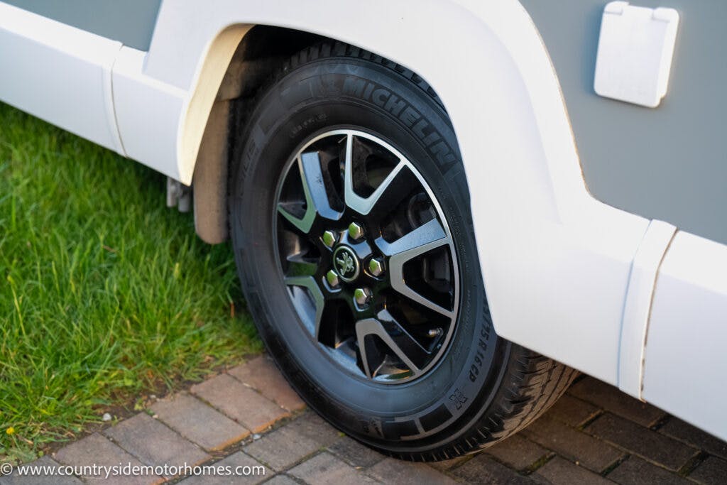 Close-up view of a 2021 Bailey Autograph 79-4i motorhome’s wheel. The tire is branded Michelin and is mounted on a black and silver alloy wheel with a branded center cap. The vehicle is parked on a paved surface with grass nearby.