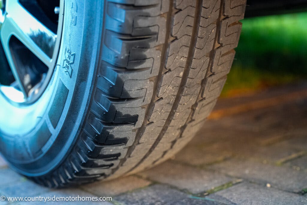 Close-up image of a vehicle tire on a brick-paved surface. The tire shows slight wear across the tread. The sidewall pattern and tread details are clearly visible. This 2021 Bailey Autograph 79-4i is part of a larger scene possibly involving a parked vehicle.