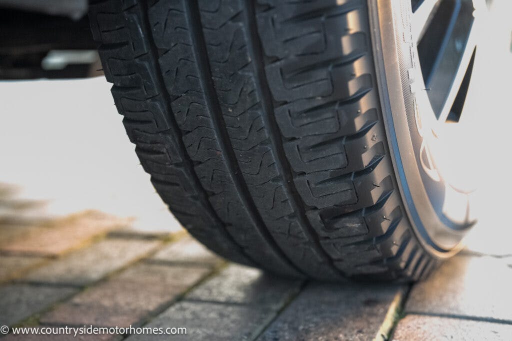 Close-up view of a car tire on a paved surface, showing the tread pattern and part of the wheel rim. The ground appears to be made of interlocking brick pavers. A 2021 Bailey Autograph 79-4i stands tall in the background. The website www.countrysidemotorhomes.com is visible in the bottom left corner.