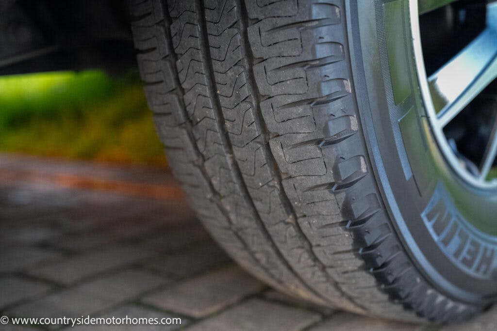 Close-up view of a car tire with a detailed tread pattern on the 2021 Bailey Autograph 79-4i. The tire is mounted on a wheel, and the background shows a paved surface and green foliage. The image focuses on the tire's surface and texture, with the website URL visible at the bottom left.