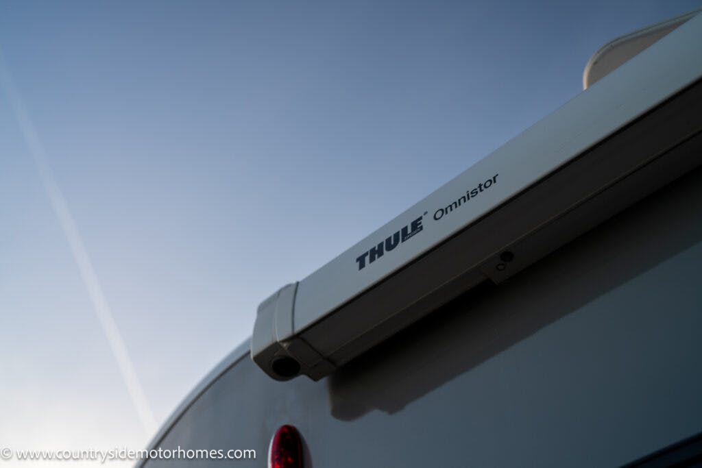 Close-up of a Thule Omnistor awning attached to the top edge of a 2021 Bailey Autograph 79-4i motorhome. The sky in the background is clear with a visible contrail. The image is watermarked with the URL "www.countrysidemotorhomes.com" in the bottom-left corner.