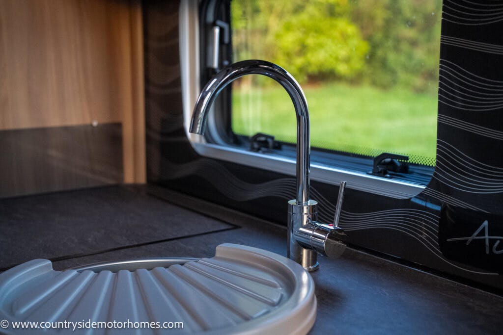 The image shows a modern kitchen sink area inside a 2021 Bailey Autograph 79-4i motorhome. There is a sleek metallic faucet next to a round sink with a removable cover. The window above the sink offers a view of green foliage outside. The website "www.countrysidemotorhomes.com" is visible in the bottom left.