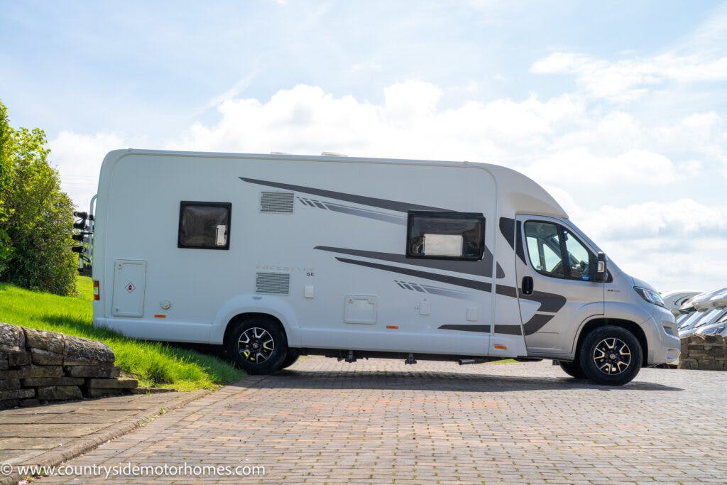 A 2019 Swift Escape 694 Freestyle motorhome parked on a paved area next to a stone wall and greenery. The vehicle features several windows and a sleek design with grey accents. The sky above is partly cloudy.