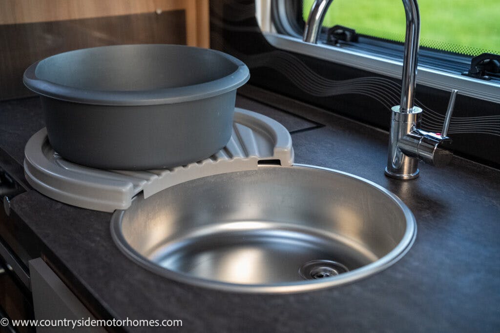 A photo of a stainless-steel sink with a chrome faucet in the kitchen of a 2021 Bailey Autograph 79-4i motorhome. A grey plastic basin is placed on a circular dish drainer next to the sink. The black countertop is visible, and a window with a partial view of greenery is in the background.