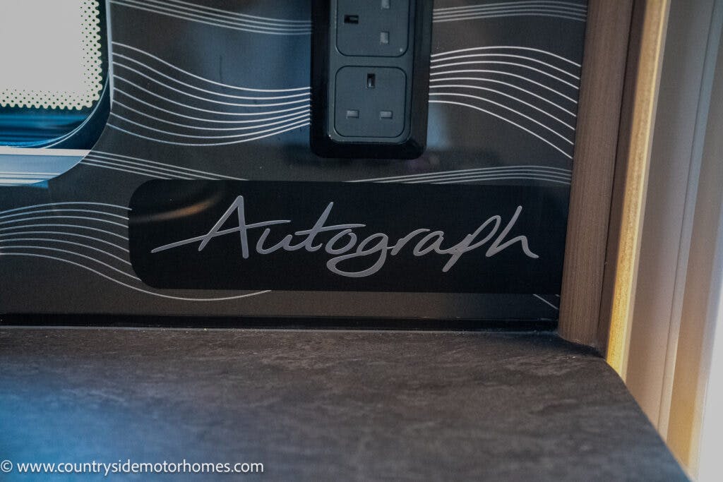 Close-up of a black and gray countertop area featuring a wall power outlet and a decorative background with white lines. The word "Autograph" is prominently displayed in cursive font, hinting at the sleek design of the 2021 Bailey Autograph 79-4i. Website, www.countrysidemotorhomes.com, is visible at the bottom left corner.