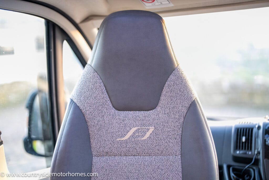 A close-up view of the driver seat in the 2021 Bailey Autograph 79-4i motorhome, featuring gray upholstery and a white embroidered logo. The interior is blurred in the background, with the website www.countrysidemotorhomes.com visible in the bottom left corner.