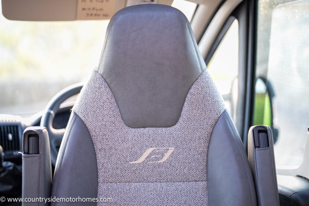 Close-up of the driver's seat in a 2021 Bailey Autograph 79-4i van. The seat is upholstered in grey fabric with a light grey patterned insert and features white stitching forming a logo in the center. The steering wheel and part of the dashboard are visible in the background.