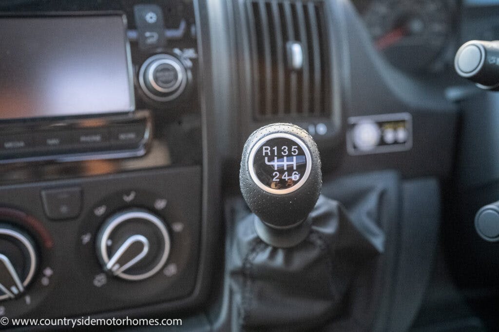 Close-up view of a manual gear shift knob in a 2021 Bailey Autograph 79-4i. The shift knob displays a gear pattern labeled R, 1, 2, 3, 4, 5, and 6 with a separate lever for 4H. The dashboard, including various controls and buttons, is visible in the background.