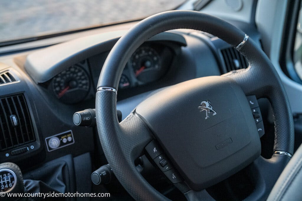 A close-up view of a steering wheel inside a Peugeot vehicle, reminiscent of the 2021 Bailey Autograph 79-4i. The dashboard and instrument panel are visible in the background, displaying speedometer and odometer. A gear stick can be seen to the left. The image includes a watermark with the website address.