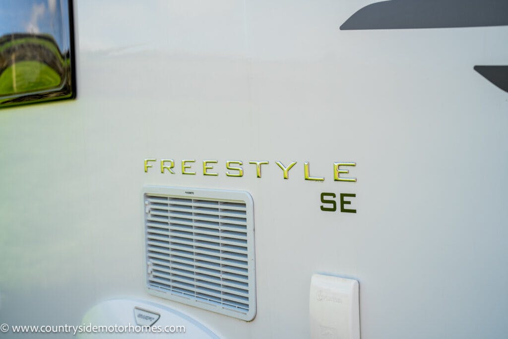 Close-up of a white 2019 Swift Escape 694 Freestyle motorhome labeled "FREESTYLE SE" with a ventilation grille below the text. The background shows a grassy area. The bottom left corner features the website www.countrysidemotorhomes.com.