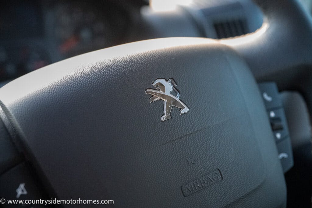 Close-up view of a Peugeot car steering wheel. The image prominently features the manufacturer's lion emblem in the center along with the word "AIRBAG" below it. In this 2021 Bailey Autograph 79-4i model, the dashboard and controls are slightly blurred in the background.
