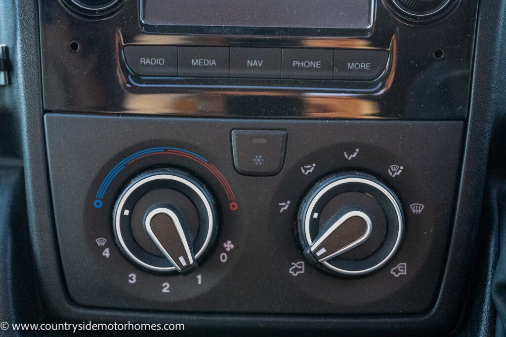 The dashboard controls of the 2021 Bailey Autograph 79-4i feature knobs for AC temperature and fan speed adjustment. The left knob adjusts temperature with blue and red indicators, while the right knob manages fan speed, defroster, and airflow direction settings.
