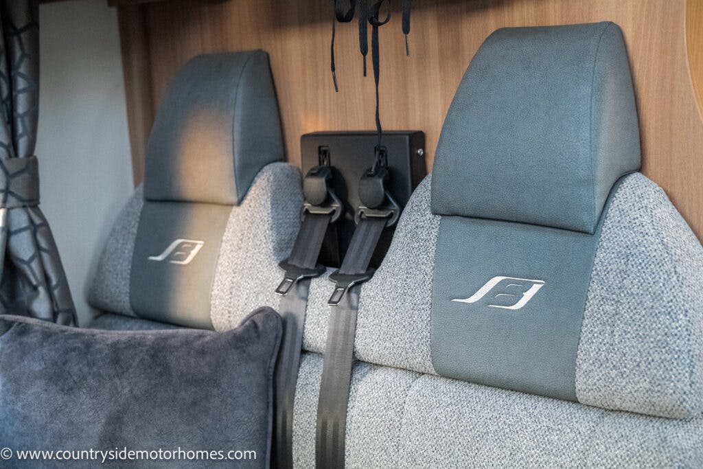 Two grey and white cushioned motorhome seats with seatbelts face forward. Both seats have an embroidered "S" logo on the headrests. The background of the 2021 Bailey Autograph 79-4i features a wooden panel and a partially visible curtain to the left.