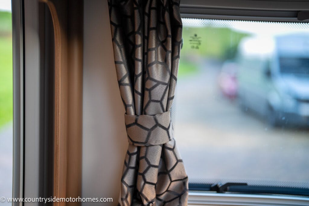 A close-up of a curtain tied back inside a 2021 Bailey Autograph 79-4i motorhome. The curtain has a geometric pattern, and the view through the window shows a parked vehicle and some greenery in the background, highlighting the cozy interior of this modern motorhome.